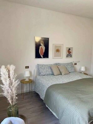 A bedroom with a bed, two bedside tables, a lamp, a vase with pampas grass, and three wall art pieces.