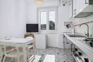 Eine kompakte Küche mit weißen Schränken, einer gefliesten Spritzschutzwand, einem kleinen Fenster und einem Fernseher.