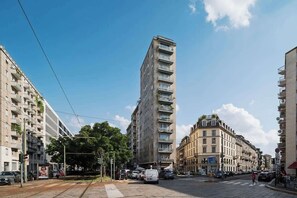 Ein mehrstöckiges Gebäude mit Balkonen, einer Straßenbahnlinie und parkenden Autos auf einer Stadtstraße.