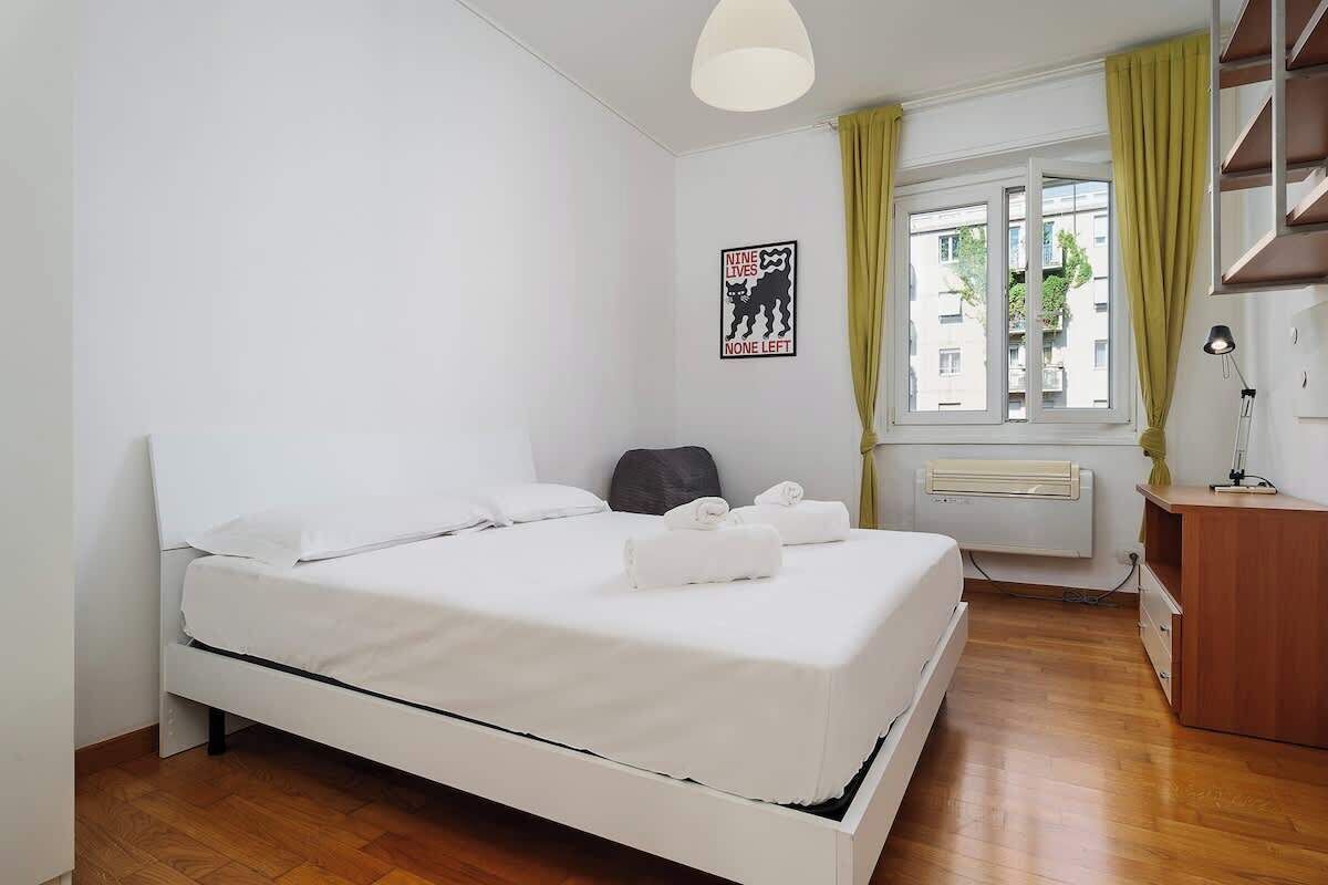A neatly made bed with white linens and towels, a window with curtains, a radiator, and a desk with a lamp.