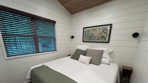 Un dormitorio con una cama, una ventana con persianas, una obra de arte enmarcada y suelo de madera.