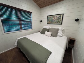 A bedroom with a bed, a window with blinds, a framed artwork, and a wooden floor.