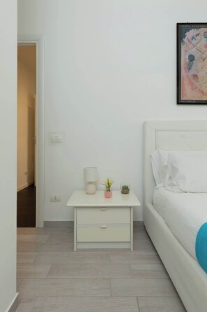 A minimalist bedroom with a white bed, a nightstand with a lamp, a small vase with flowers, and a framed artwork on the wall. - Holi Rome - Bright&Comfy apt inTrastevere (Rome)