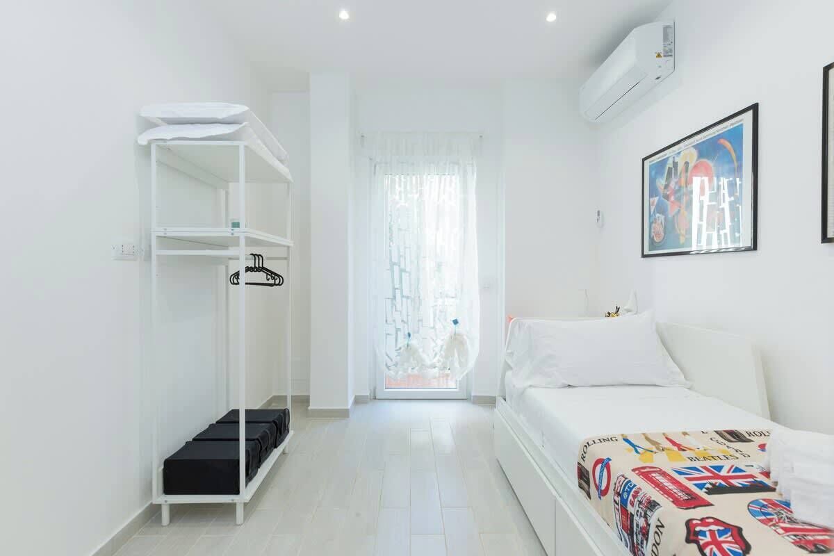 Una camera da letto con un letto, un armadio, una mensola con borse nere, una finestra con tende e un condizionatore d'aria a parete.