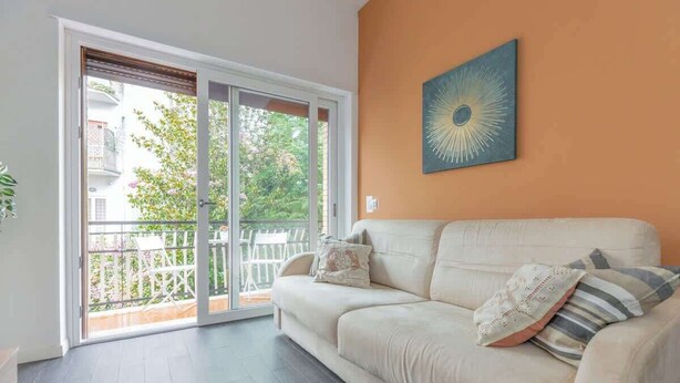 A living room with a beige sofa, a large window, and a sliding glass door leading to an outdoor balcony. - Holi Rome - Bright&Comfy apt inTrastevere (Rome)
