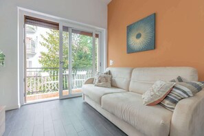 A living room with a beige sofa, a large window, and a sliding glass door leading to an outdoor balcony. - Holi Rome - Bright&Comfy apt inTrastevere (Rome)