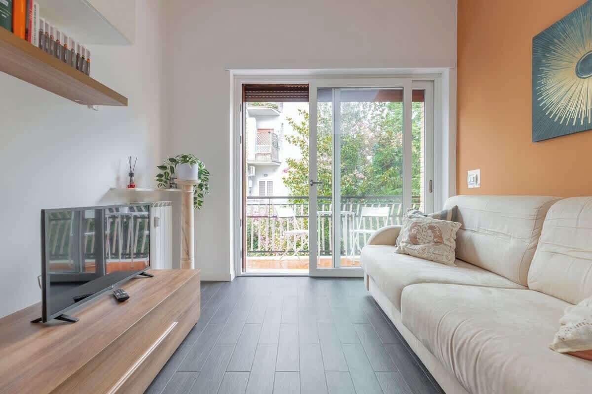Een moderne woonkamer met een bank, een televisie, een schuifdeur naar een balkon en een houten plank met boeken.