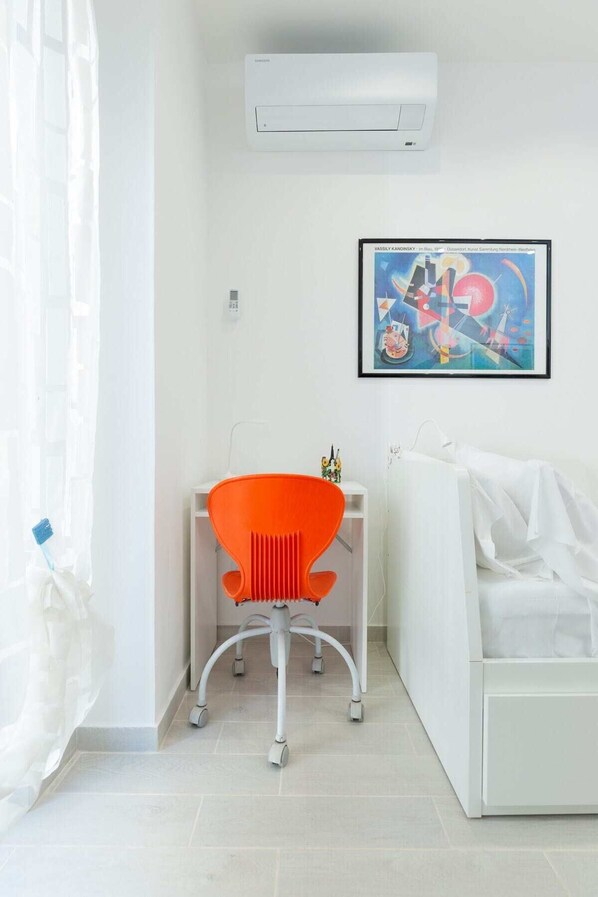 Una habitación con un equipo de aire acondicionado blanco, una obra de arte enmarcada en la pared, un sofá blanco y una silla naranja brillante.
