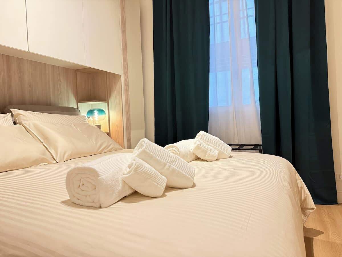 Ein ordentlich bezogenes Bett mit aufgerollten Handtüchern, einer Nachttischlampe und einem Fenster mit Vorhängen.