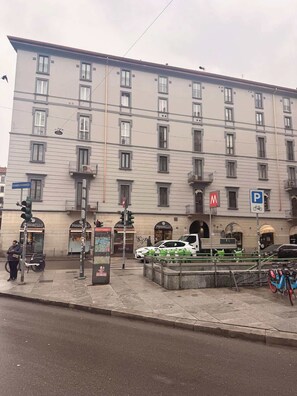 Un immeuble à plusieurs étages, avec une série de boutiques au rez-de-chaussée, un panneau indiquant un parking et un passage piéton.