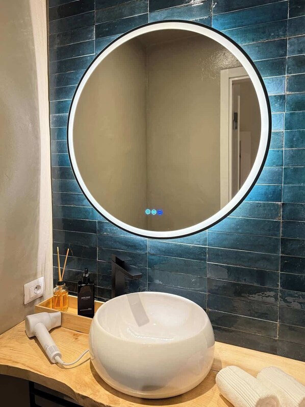 Une salle de bain avec un miroir rond, un lavabo sur colonne blanc et un sèche-cheveux posé sur le comptoir.