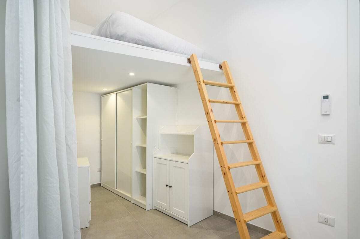 A room with a loft bed, built-in shelving, a ladder, and a white wardrobe.