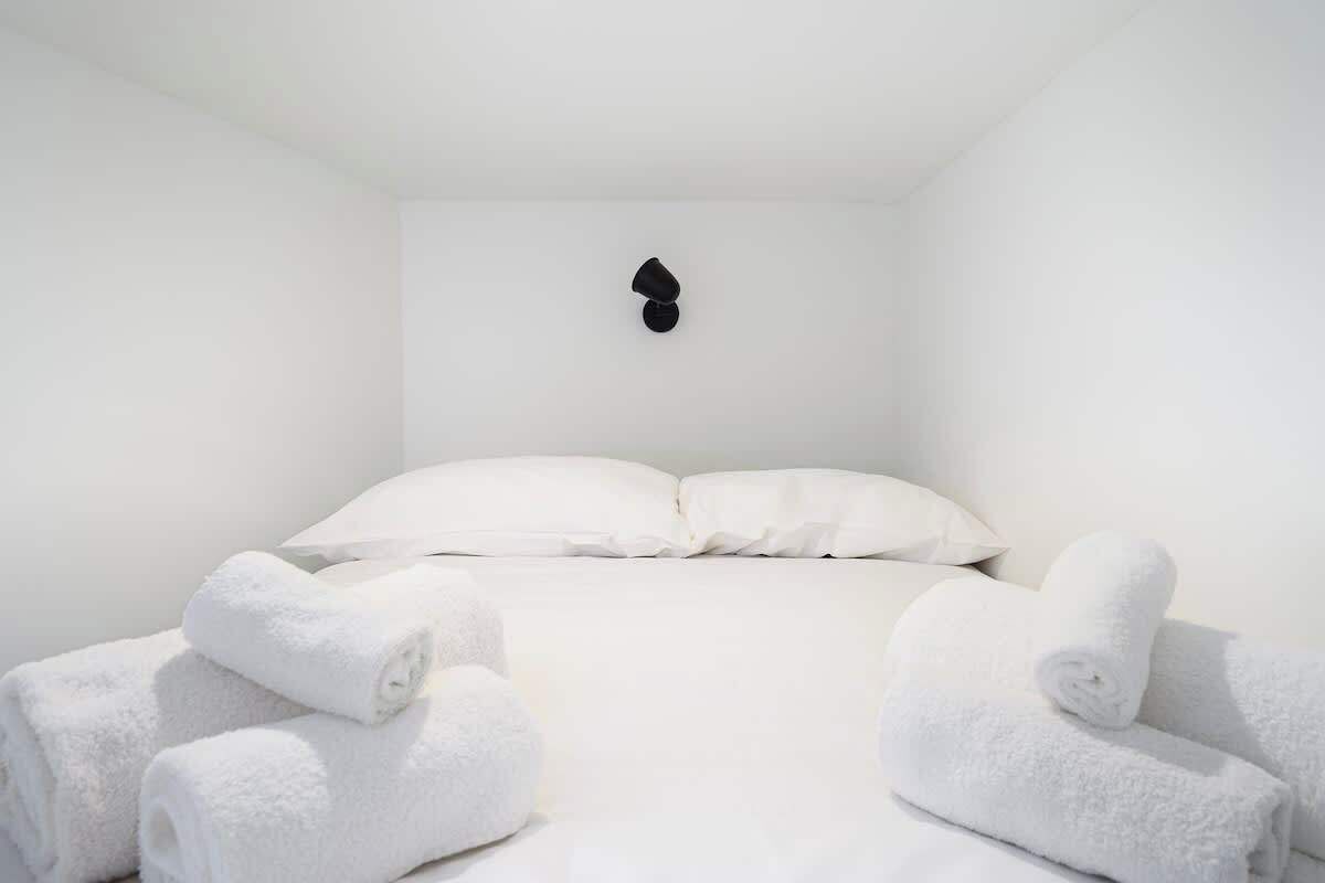 A minimalist hotel room with a neatly made bed, white linens, and a single black wall-mounted light fixture.