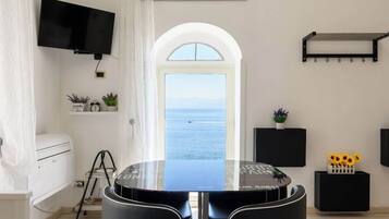 Une chambre moderne avec une table ronde, un téléviseur à écran plat, une vue sur la mer par une porte en arc de cercle et une petite étagère avec des plantes.