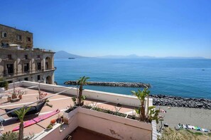 Eine Dachterrasse mit Meerblick, ein Gebäude mit Bögen und eine felsige Küstenlinie.