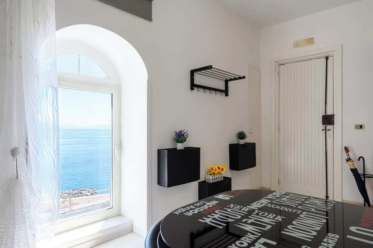 Una stanza con vista sul mare, un tavolo rotondo con piano nero e mensole nere a parete con piccole piante.
