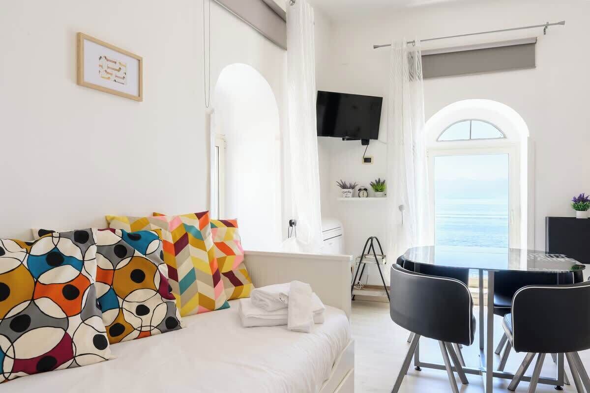 Une chambre d’hôtel moderne équipée d’un lit, d’une table à manger avec des chaises, d’un téléviseur fixé au mur et offrant une vue sur la mer par une fenêtre en arc de cercle.