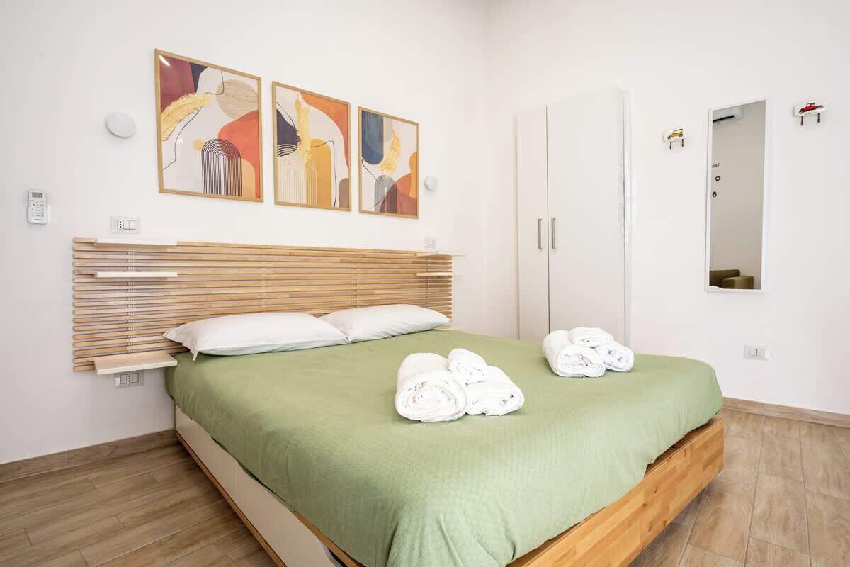 Habitación de hotel con cama, cubrecama verde, cabecera de madera, obra de arte en la pared, armario blanco y espejo.