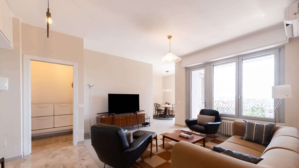 A modern living room with a sofa, armchair, TV, dining area, and large windows. - Skyline View - Design Apartment - Bicocca Metro (Milano)