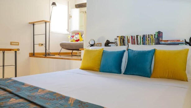 Een slaapkamer met een bed, kussens, een bureau, een stoel, een lamp en een boekenplank vol boeken.
