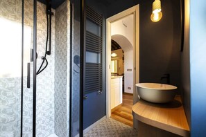 A modern bathroom with a glass shower enclosure, a wooden vanity with a large vessel sink, and a door leading to a kitchen area. - Monti - Stunning Loft in the Heart of Rome's Monti (Rome)