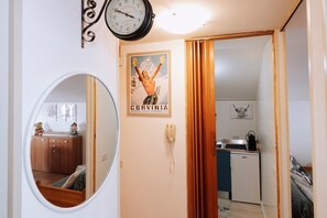 Una habitación con un espejo redondo, un reloj, un cartel, un teléfono y una puerta que da a una cocina.