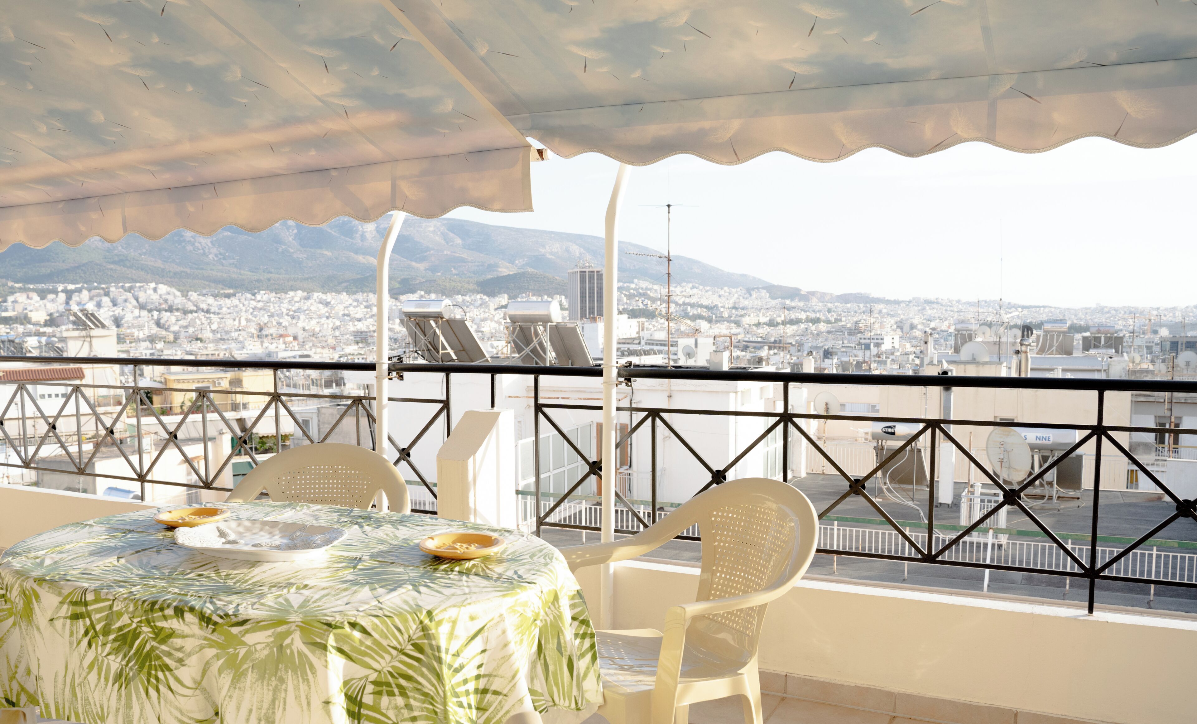 A balcony with a table and chairs, overlooking a cityscape with mountains in the distance.