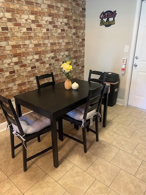 A dining table with chairs, a vase with flowers, and a brick wall. - Better Days (Charlotte)