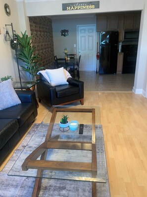 A living room with a black leather sofa, a glass coffee table with a plant and a candle, and a dining area with a table and chairs. - Better Days (Charlotte)