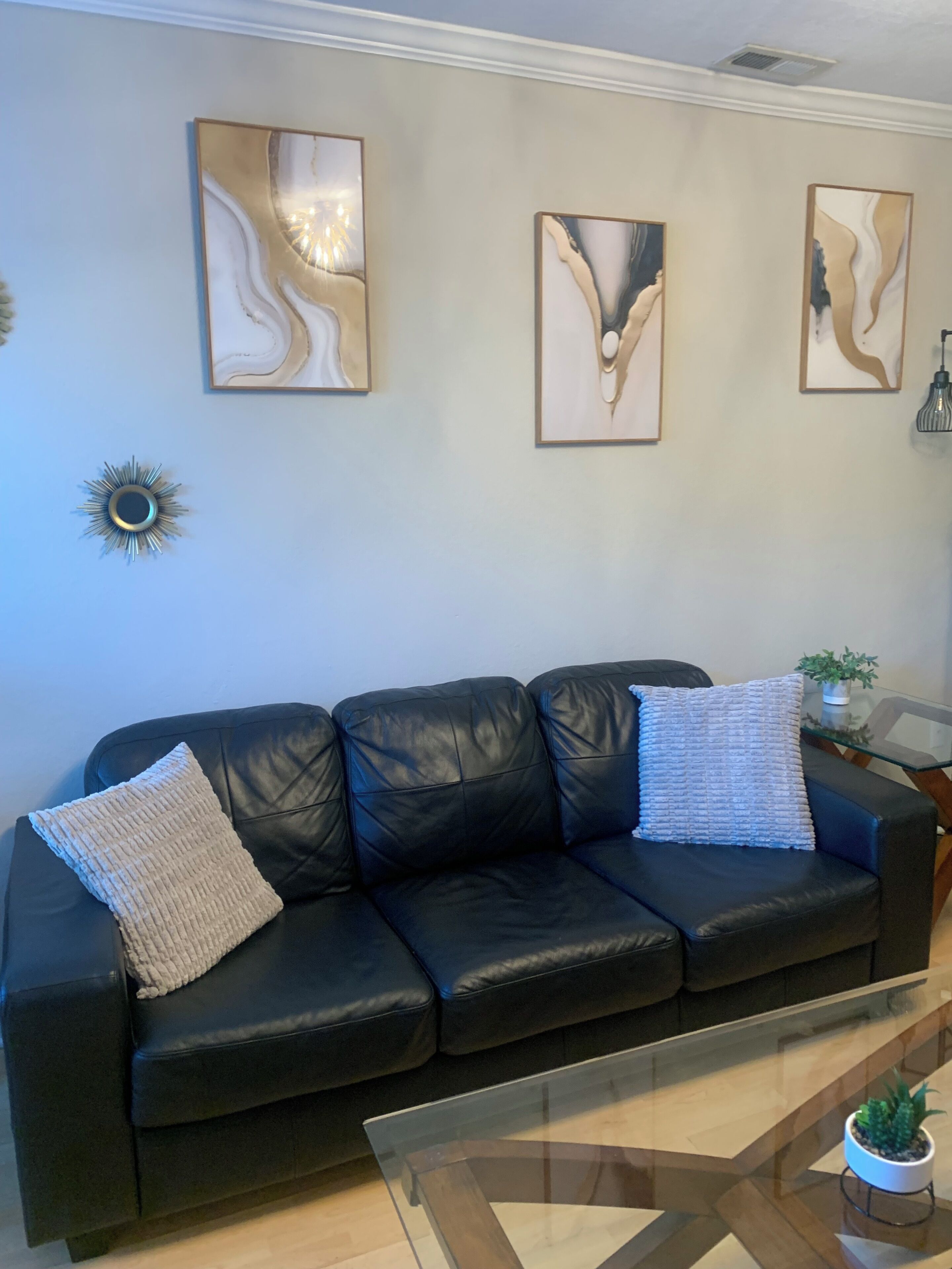 A black leather sofa with two pillows, a glass coffee table, and abstract wall art.