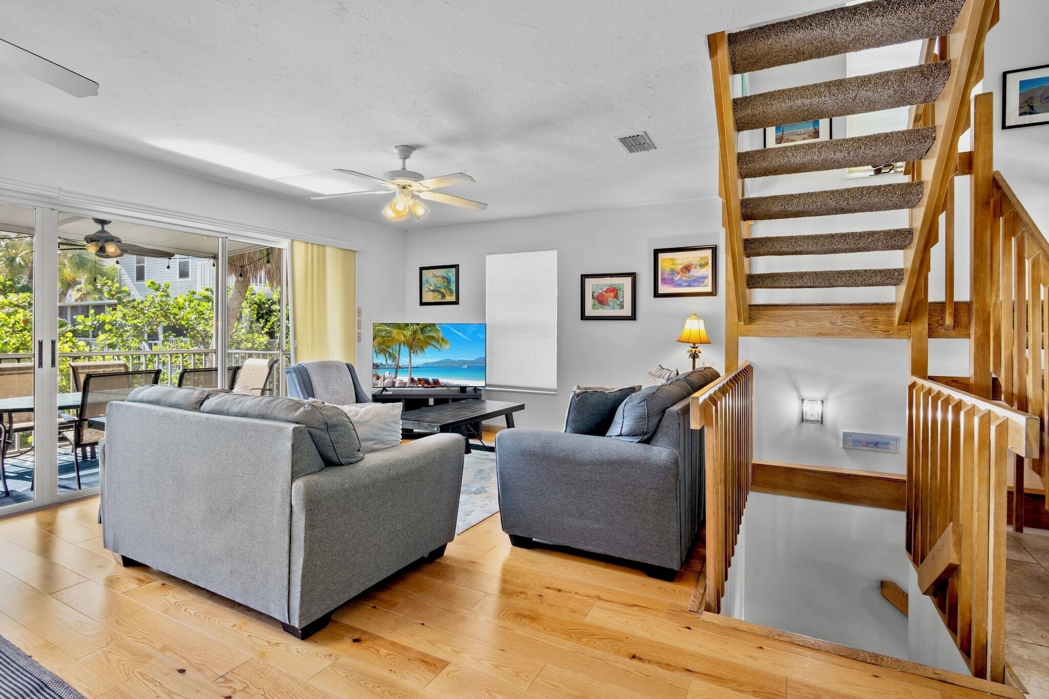 A modern living room with a wooden staircase, grey sofas, a TV, and a ceiling fan.