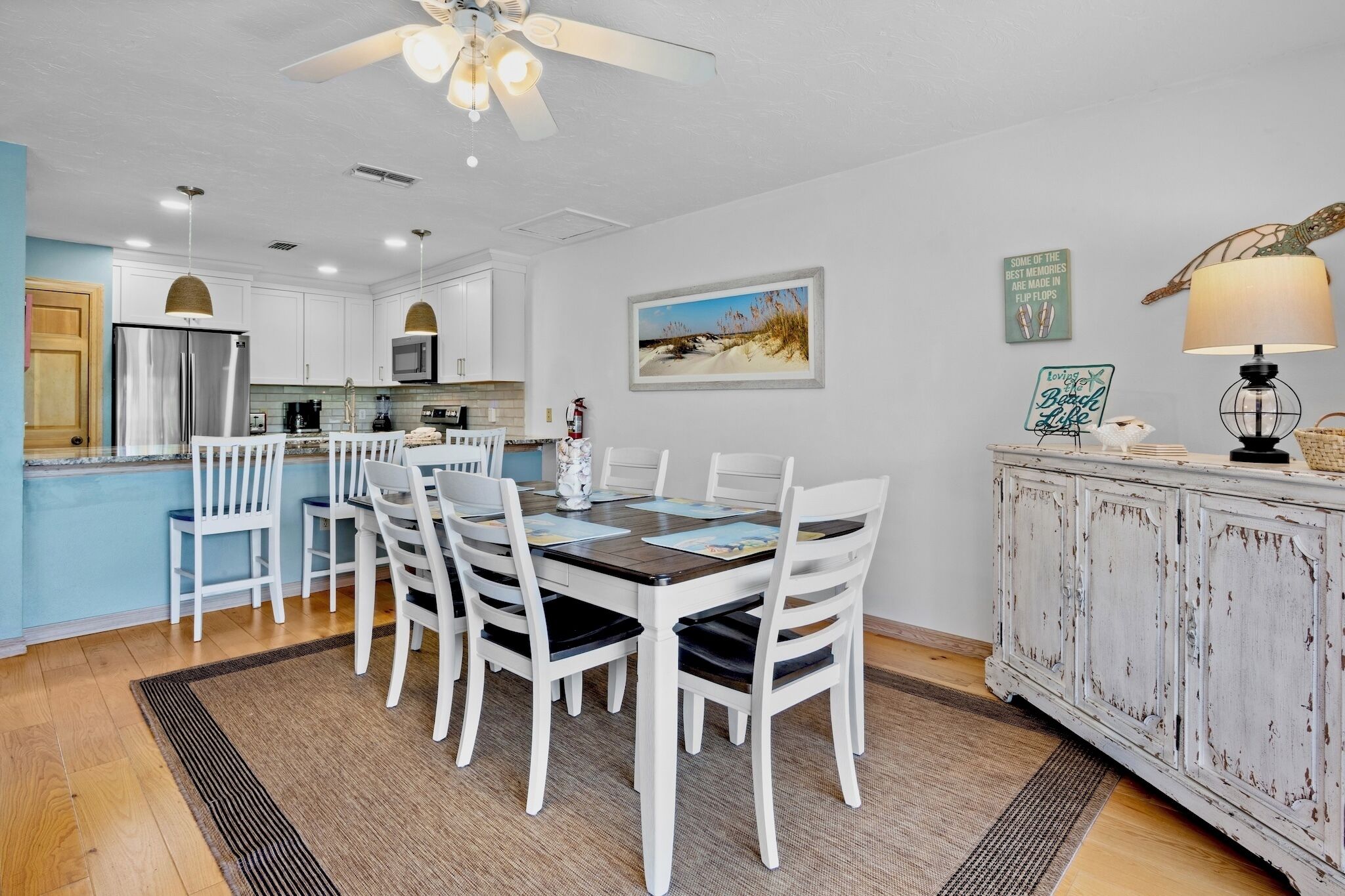 A dining area with a white table and chairs, a kitchen in the background, and a decorative lamp on a cabinet.