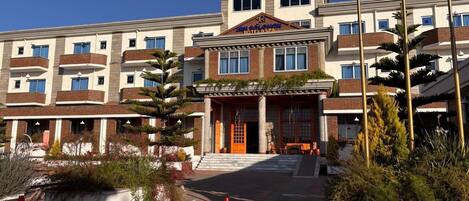 A multi-story hotel with a central entrance, balconies, and a landscaped garden area.