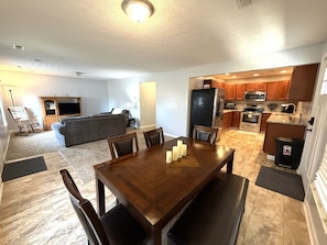 A dining area with a wooden table, chairs, and candles. There is a kitchen with modern appliances and wooden cabinets.