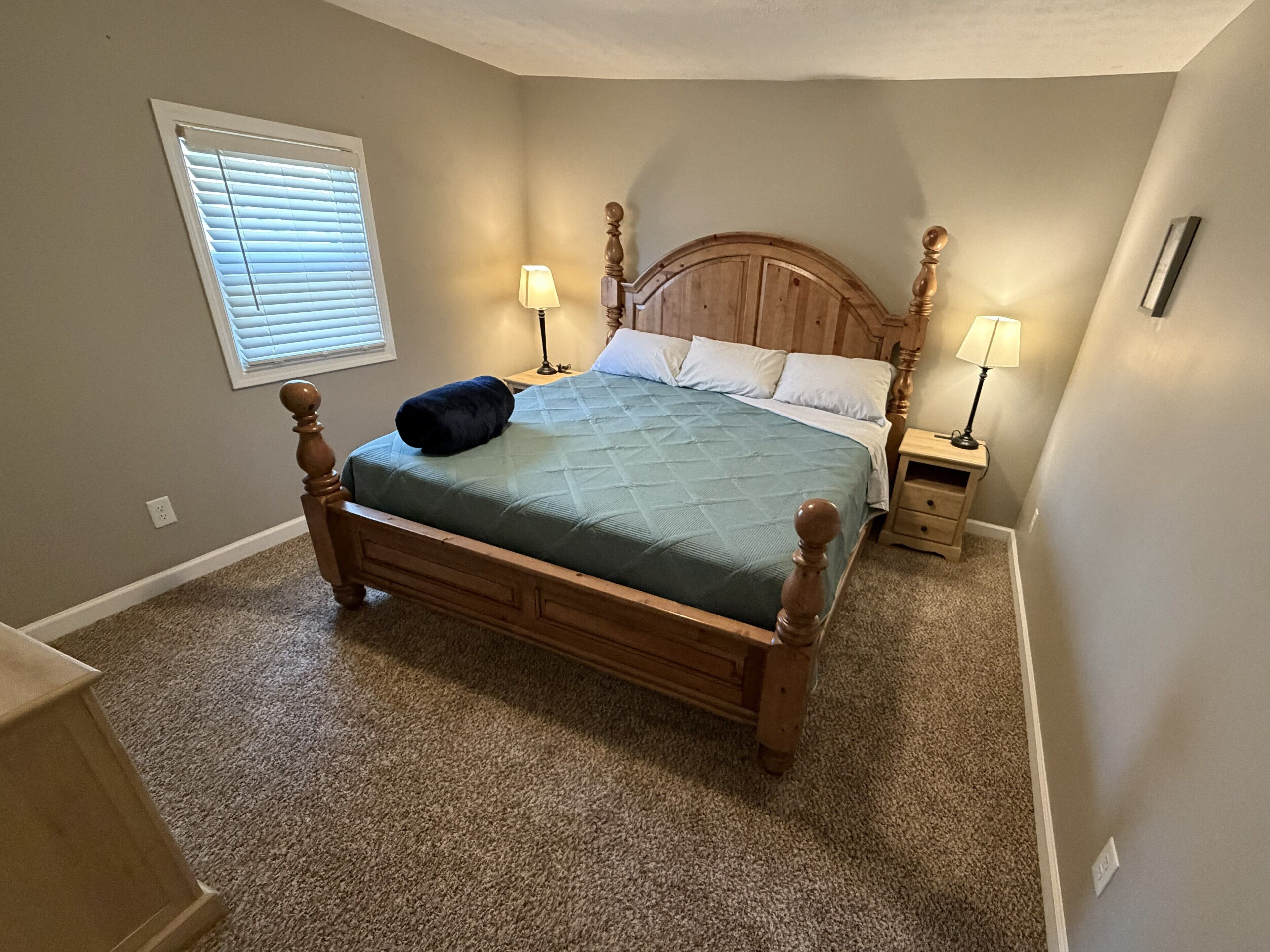 A bedroom with a wooden bed, two bedside tables with lamps, a window with blinds, and a carpeted floor.