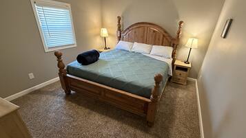A bedroom with a wooden bed, two bedside tables with lamps, a window with blinds, and a carpeted floor.