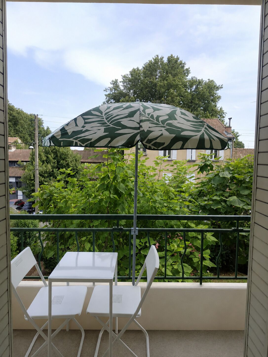 Ein Balkon mit Tisch und Stühlen, einem gemusterten Sonnenschirm und Blick auf Häuser und Bäume.