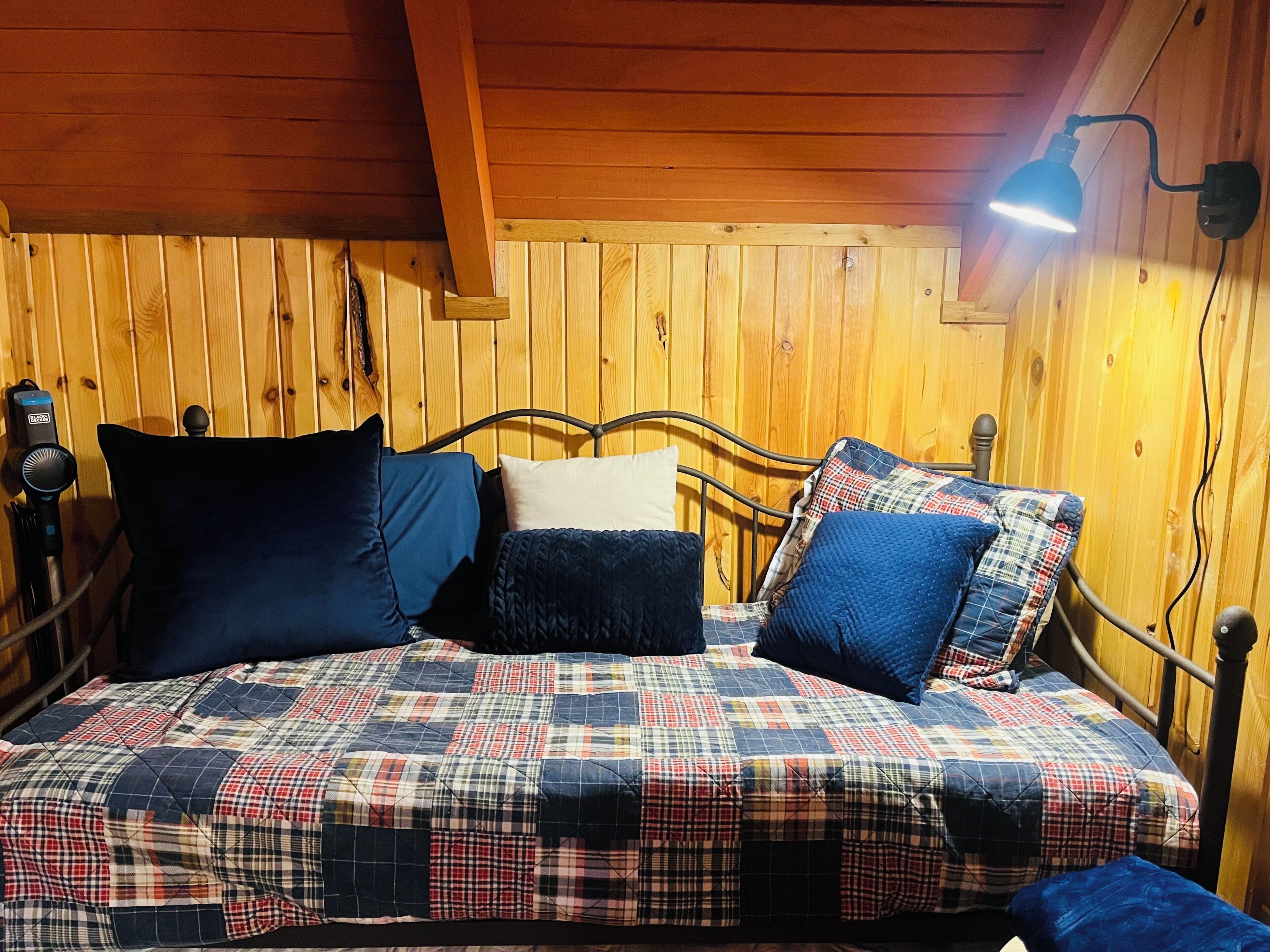 Une pièce aux murs lambrissés, avec un lit orné d’une courtepointe à carreaux, d’une tête de lit et d’une lampe fixée au mur.