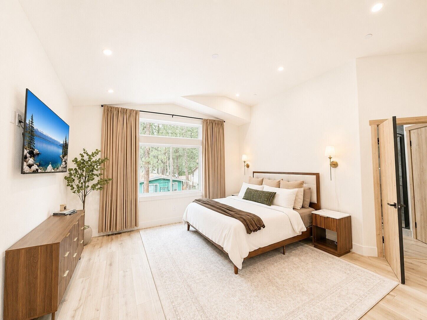 Une chambre à coucher comprenant un lit, des tables de chevet, une commode, une fenêtre avec des rideaux, un radiateur, un téléviseur fixé au mur et une porte.