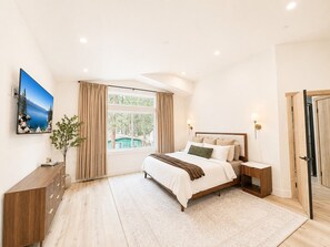 A bedroom with a bed, bedside tables, a nightstand, a window with curtains, a radiator, a wall-mounted TV, and a door.