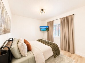 A bedroom with a bed, a TV, curtains, a painting, and a window.