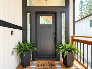 A dark front door with a welcome mat, two potted plants, and a wooden railing.