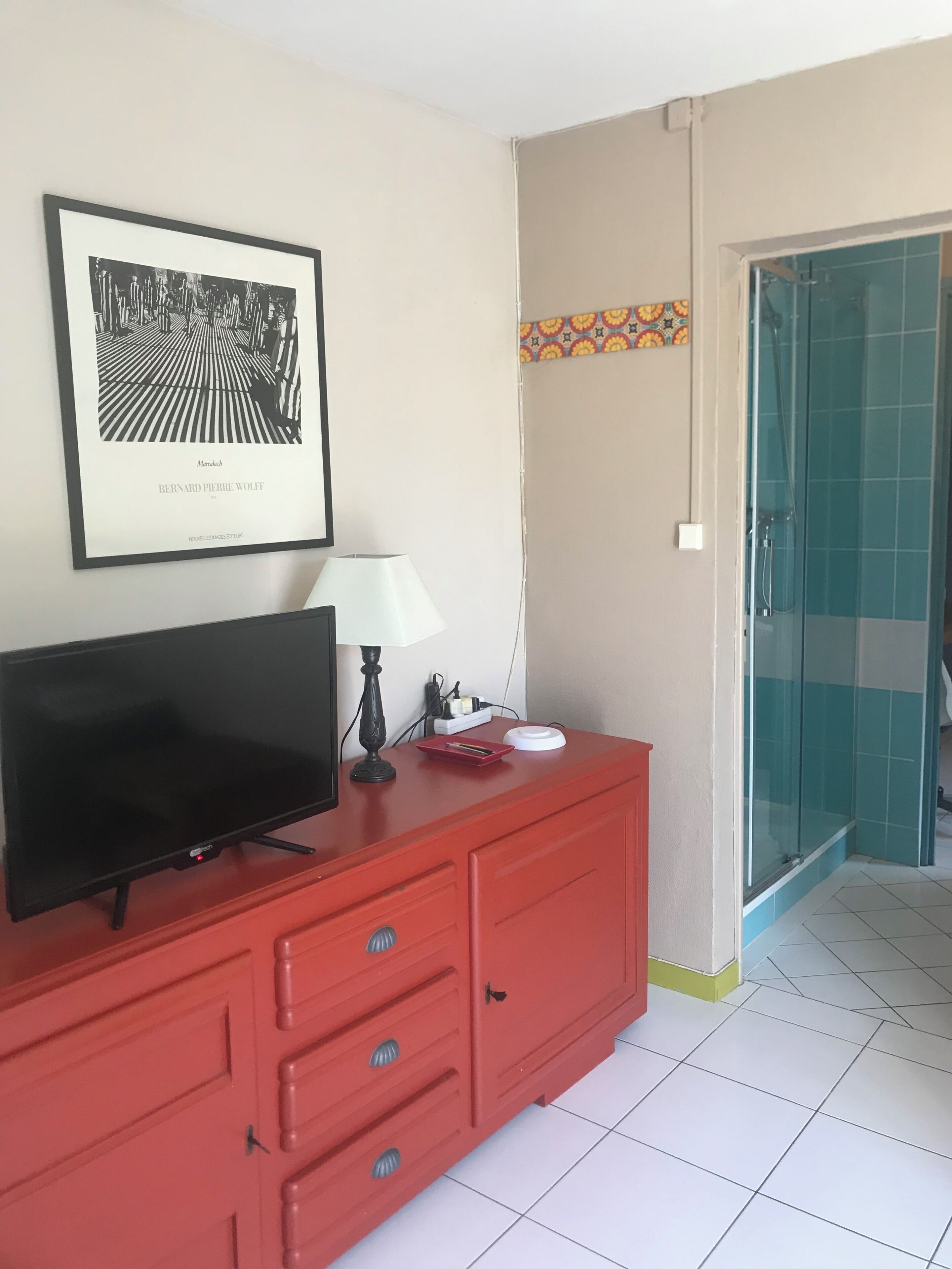 A room with a flat-screen TV on a red cabinet, a framed picture on the wall, a white lamp, and a shower area with blue tiles.