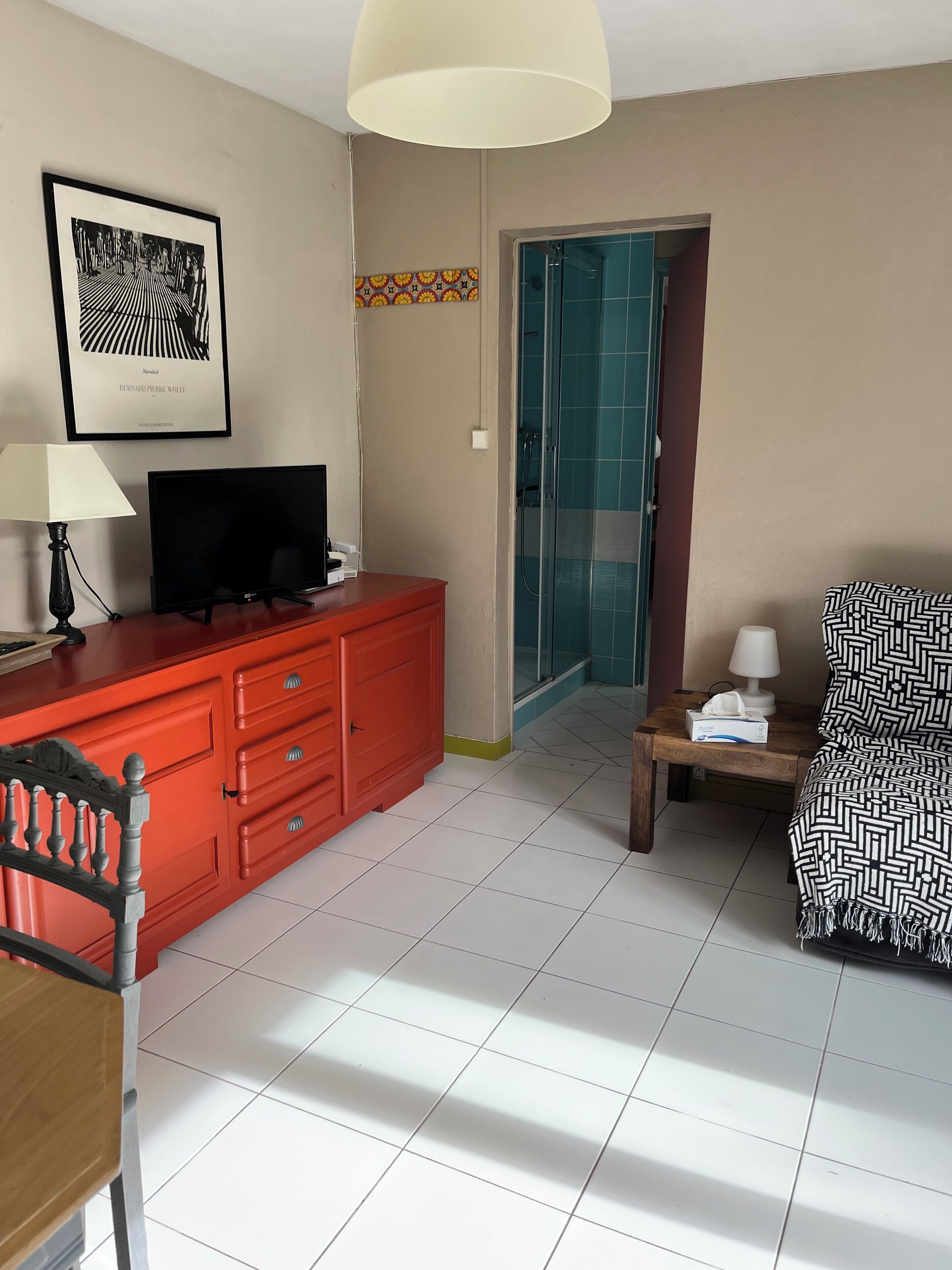 Une chambre d’hôtel avec une commode rouge, un téléviseur, un tableau encadré au mur, un lit recouvert d’une couverture à motifs et un espace douche.