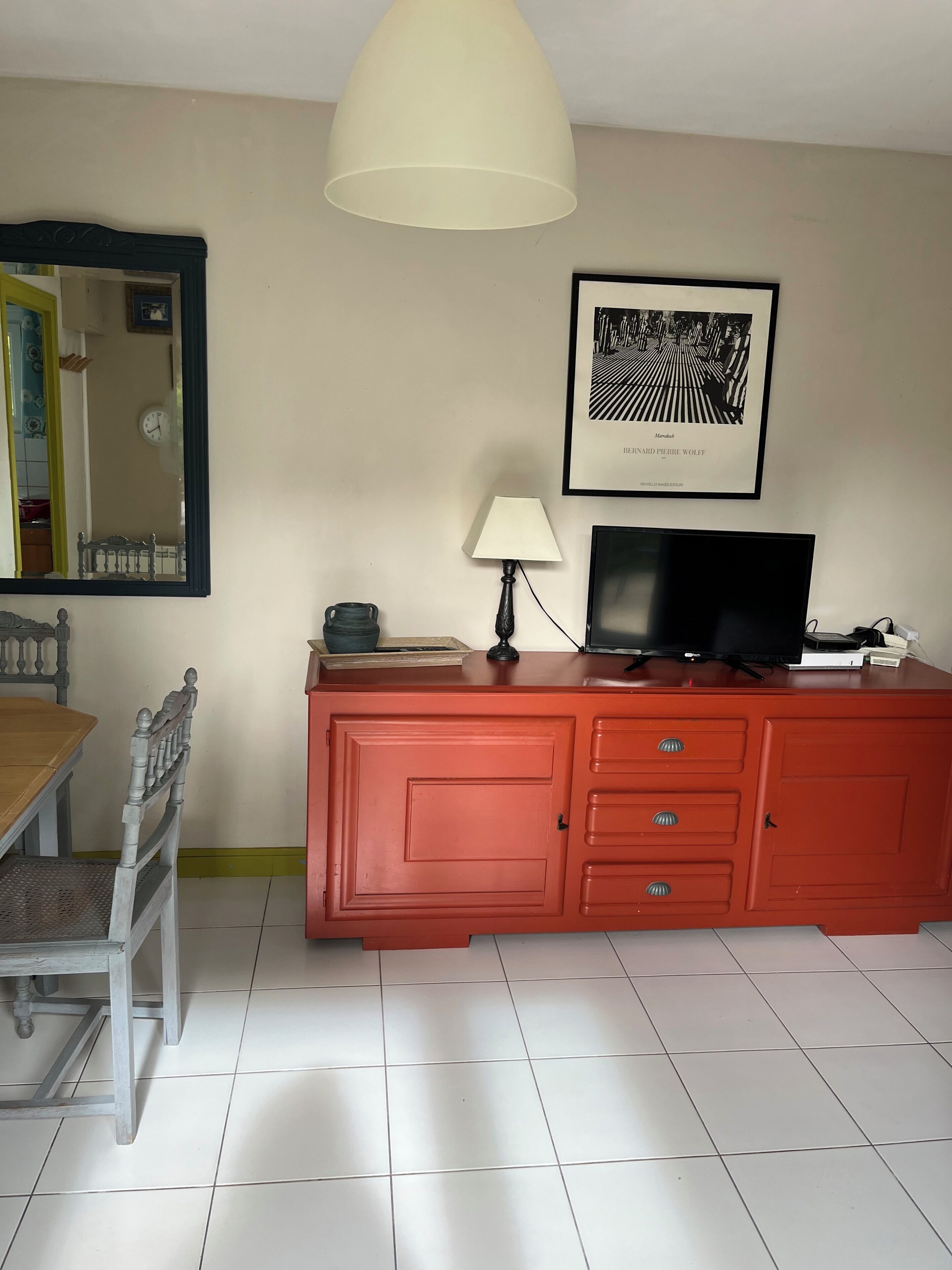 Une pièce comprenant une table à manger, un buffet rouge avec des tiroirs, un téléviseur, une lampe et un tableau encadré accroché au mur.