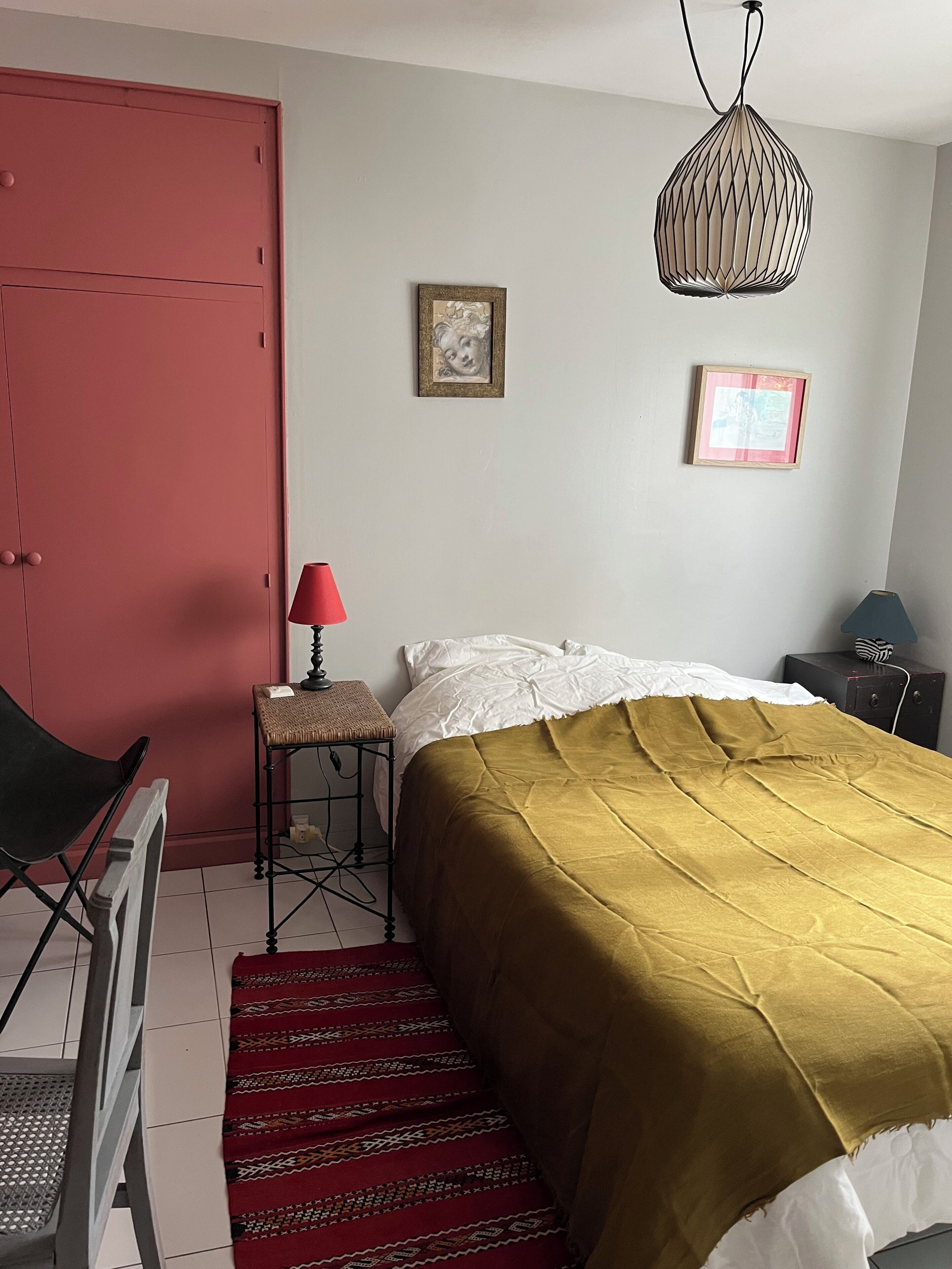 Ein Schlafzimmer mit einem Bett, einer roten Tür, einem kleinen Tisch mit Lampe und zwei gerahmten Bildern an der Wand.