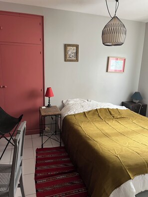 A bedroom with a bed, a red door, a small table with a lamp, and two framed pictures on the wall. - Lovely 1-bedroom house in Saint-Nazaire near the coast (Saint-Nazaire)
