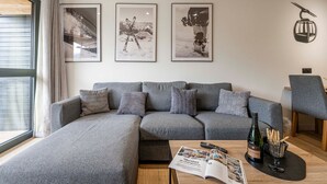 En moderne stue med en grå hjørnesofa, et sofabord med et magasin, en flaske vin og en skål med snacks. Der er billeder i rammer på væggen og en lampe til højre.
