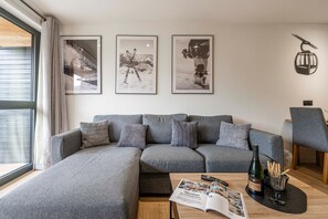 A modern living room with a grey sectional sofa, a coffee table with a magazine, a bottle of wine, and a bowl of snacks. There are framed pictures on the wall and a lamp on the right. - Suite Myrtilles - Bel appartement moderne pour 4 (Saint-Chaffrey)