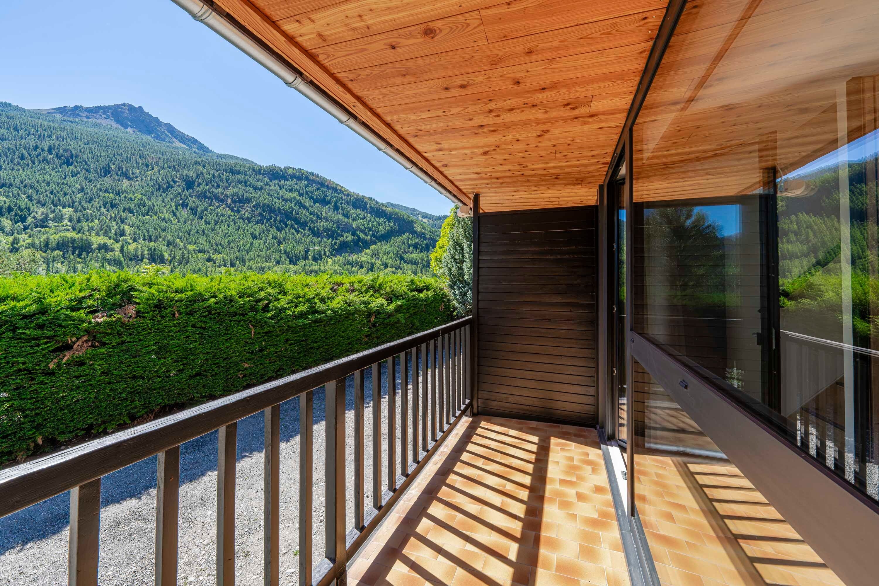 A balcony with a wooden ceiling, a metal railing, and a view of a forested mountain.